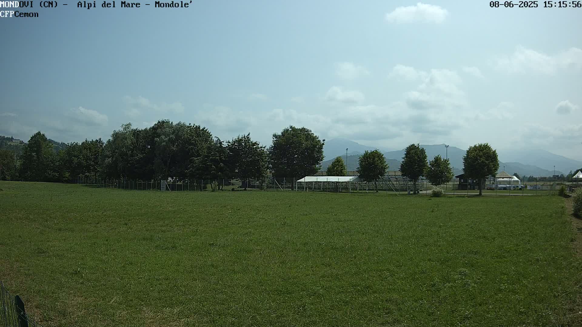 A grassy field with a line of trees and a covered structure behind a fence, under a partly cloudy sky and mountains in the distance.