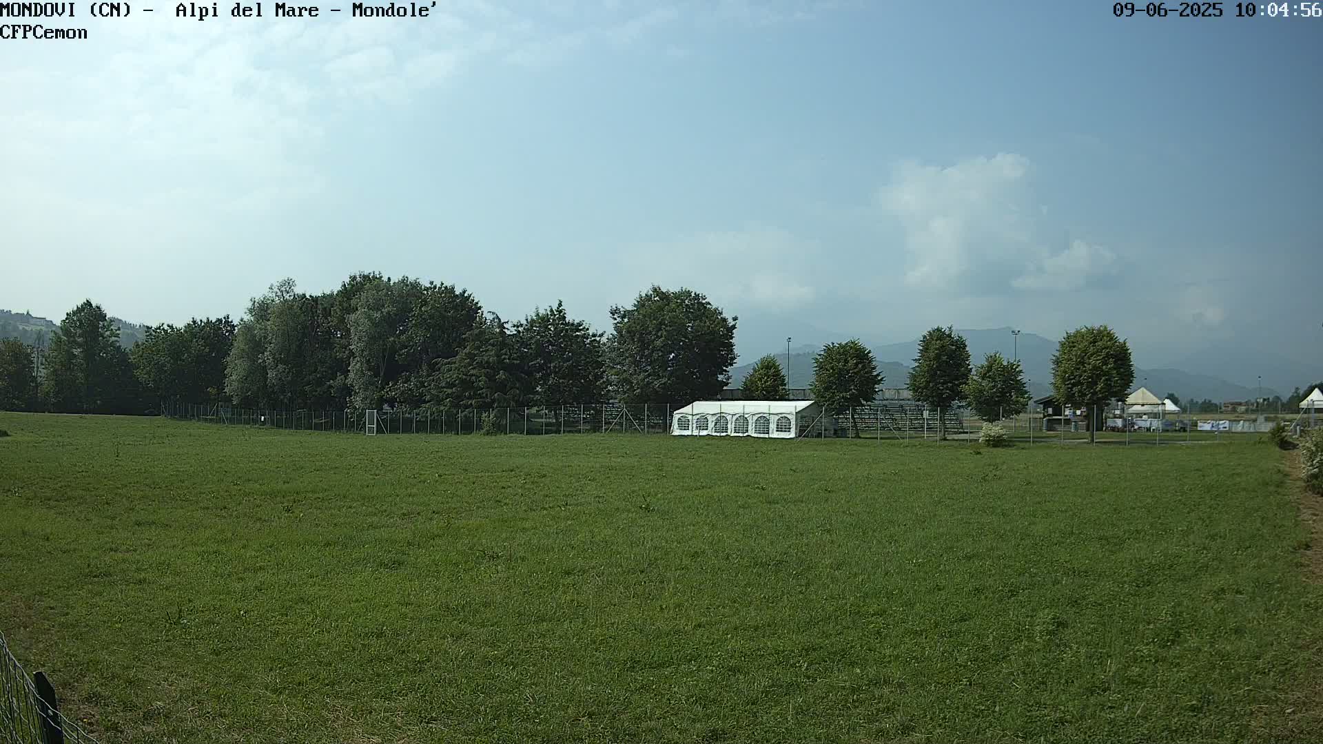 A large green field with a white tent and trees behind a fence, under a mostly clear sky.