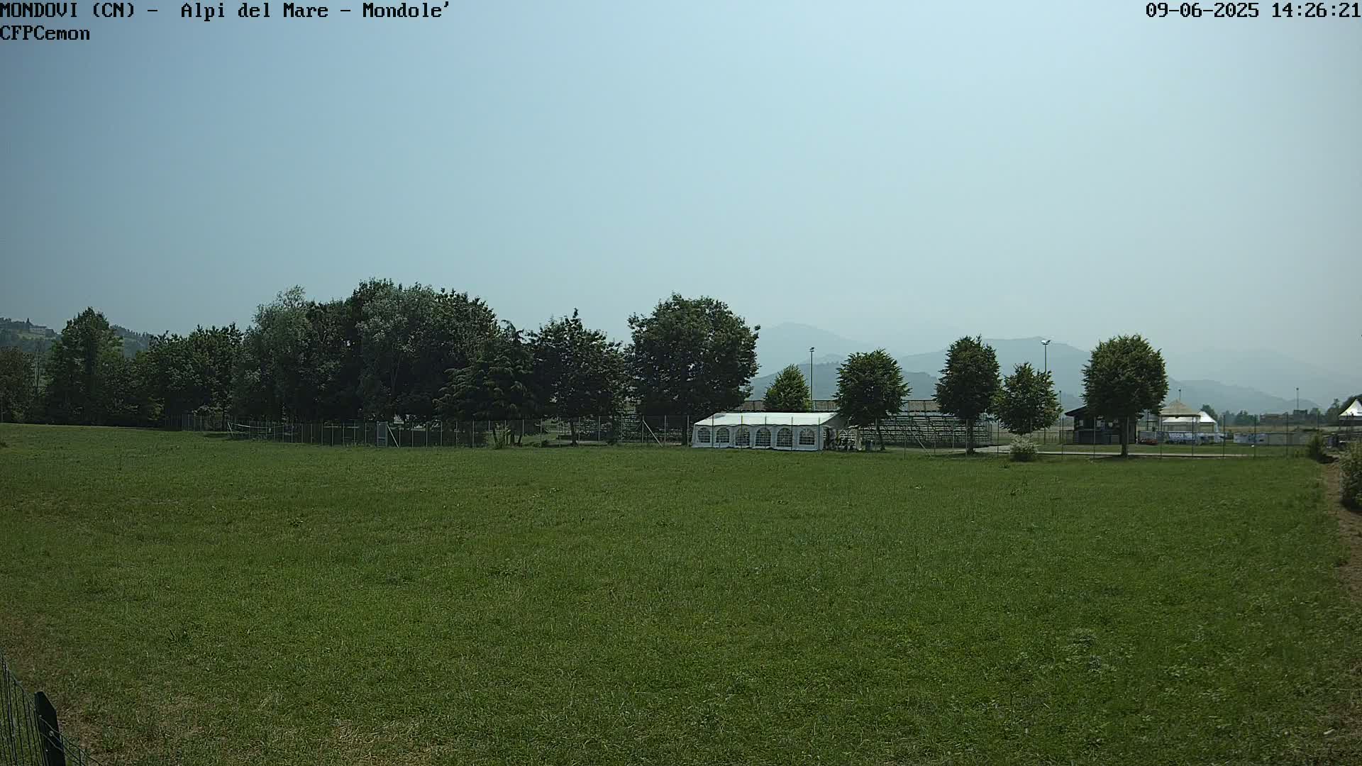A large grassy field stretches out before a line of trees and a white tent, with hazy mountains visible in the distance under a clear, light-blue sky.