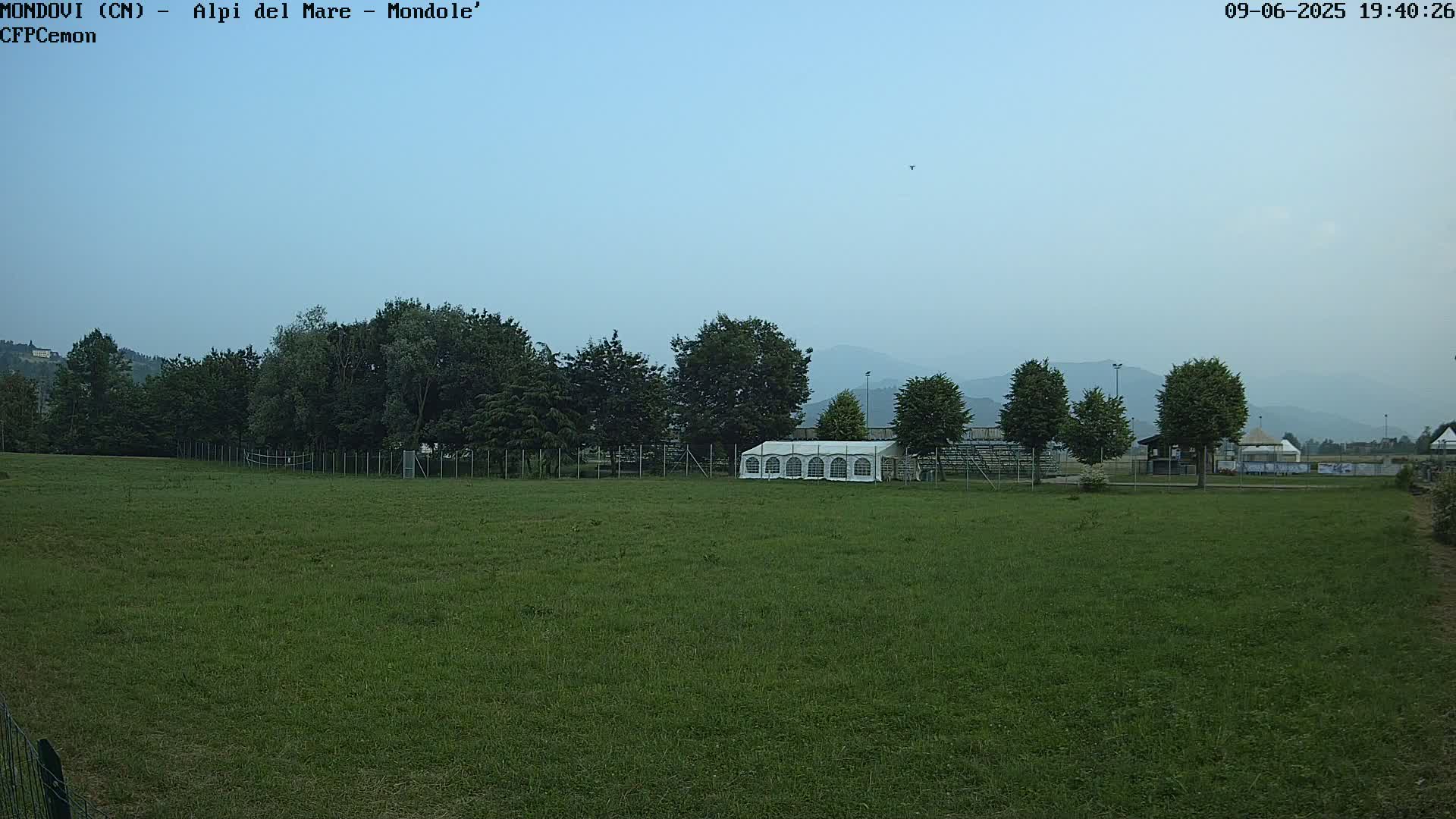 A large grassy field with a white tent and trees in the background under a hazy sky.