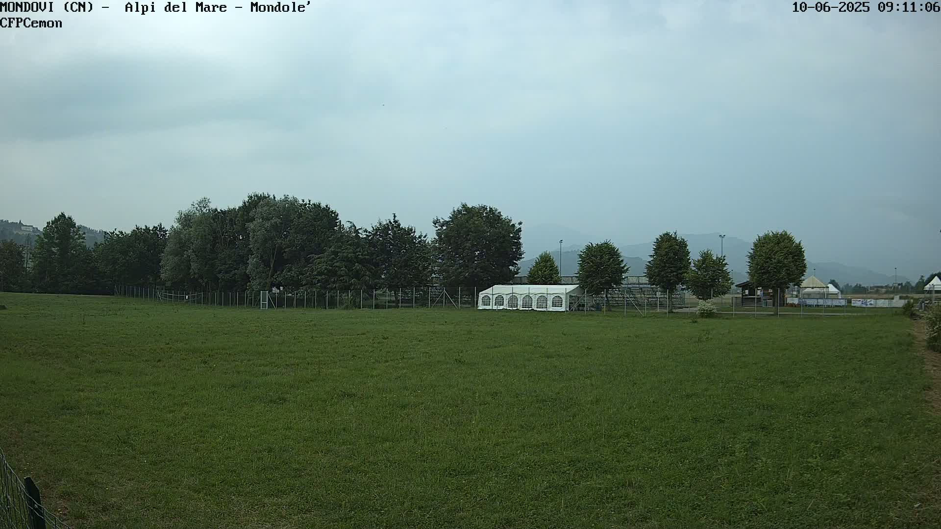 A large grassy field with a white tent and trees behind a fence, under an overcast sky.