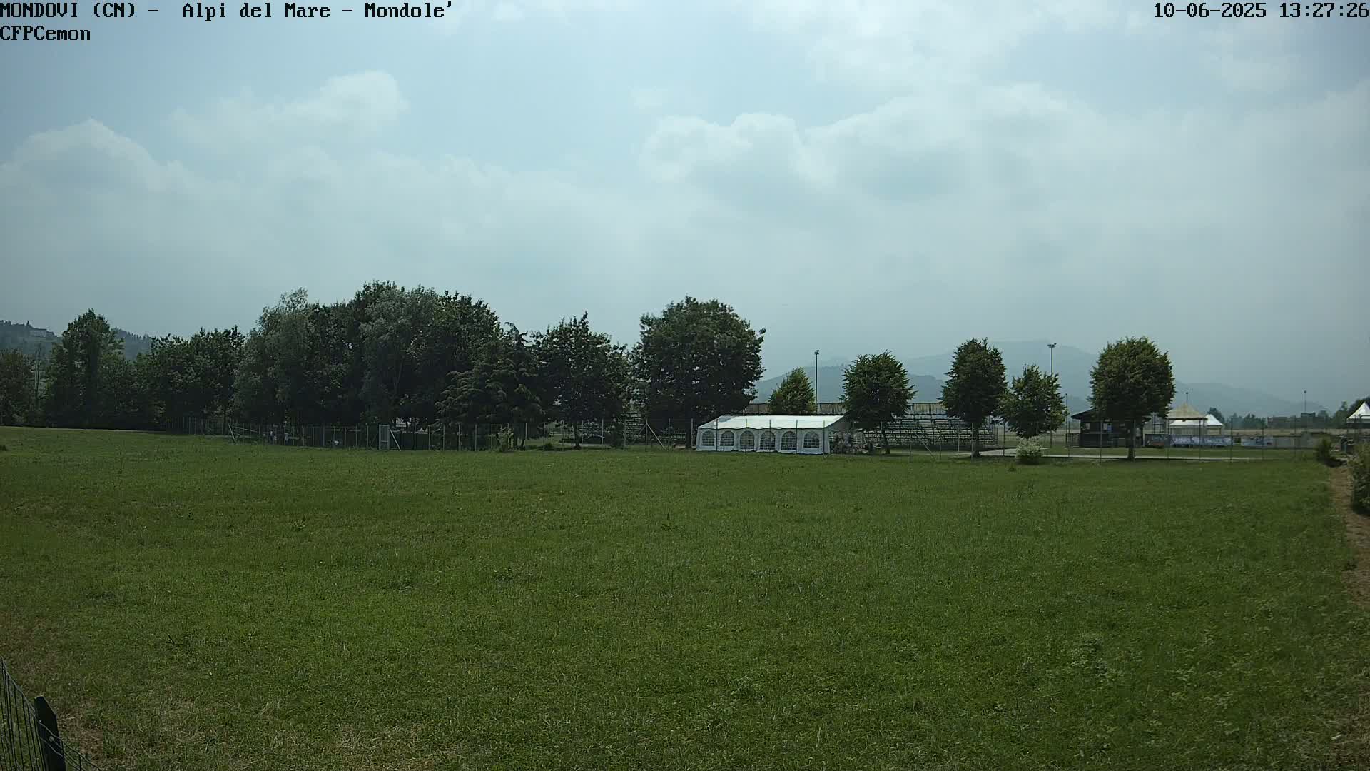 A large grassy field stretches out under a partly cloudy sky, featuring a line of trees behind a white tent near a fence.