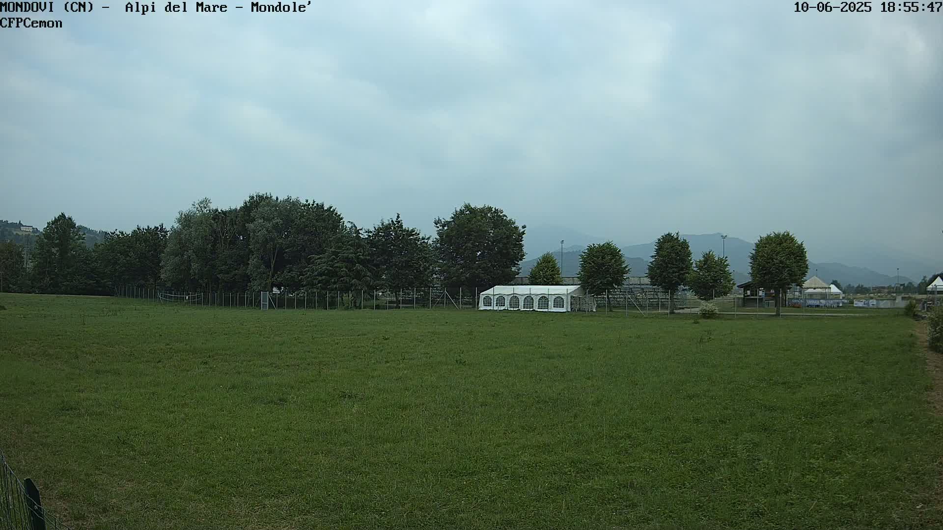 A large grassy field with a white tent sits behind a line of trees under an overcast sky, mountains visible in the distance.
