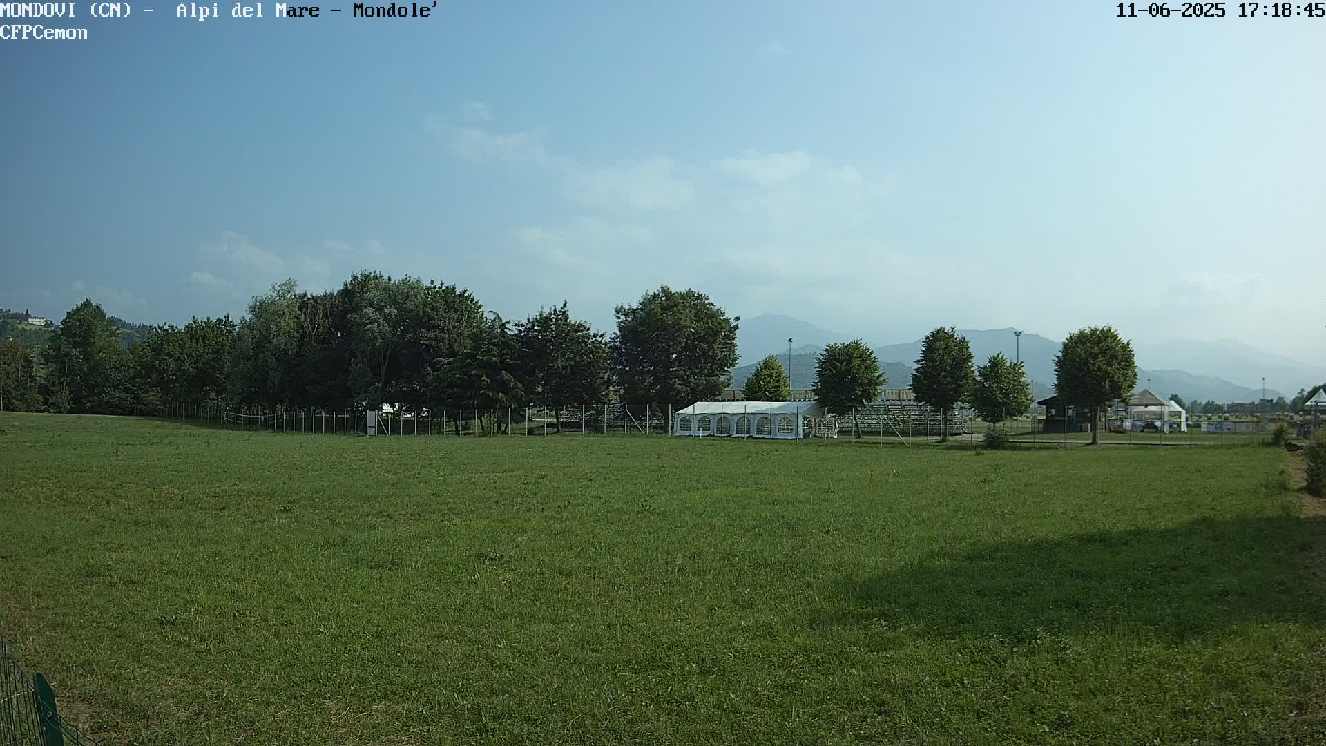 A large grassy field with a white tent and trees in the background under a mostly clear sky.