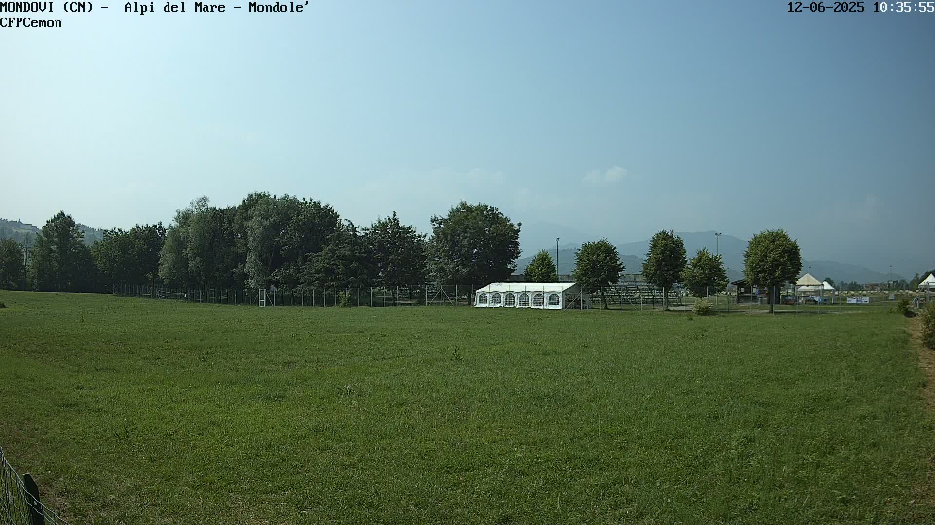 A large grassy field with a white tent, trees, and a fence, under a clear blue sky.
