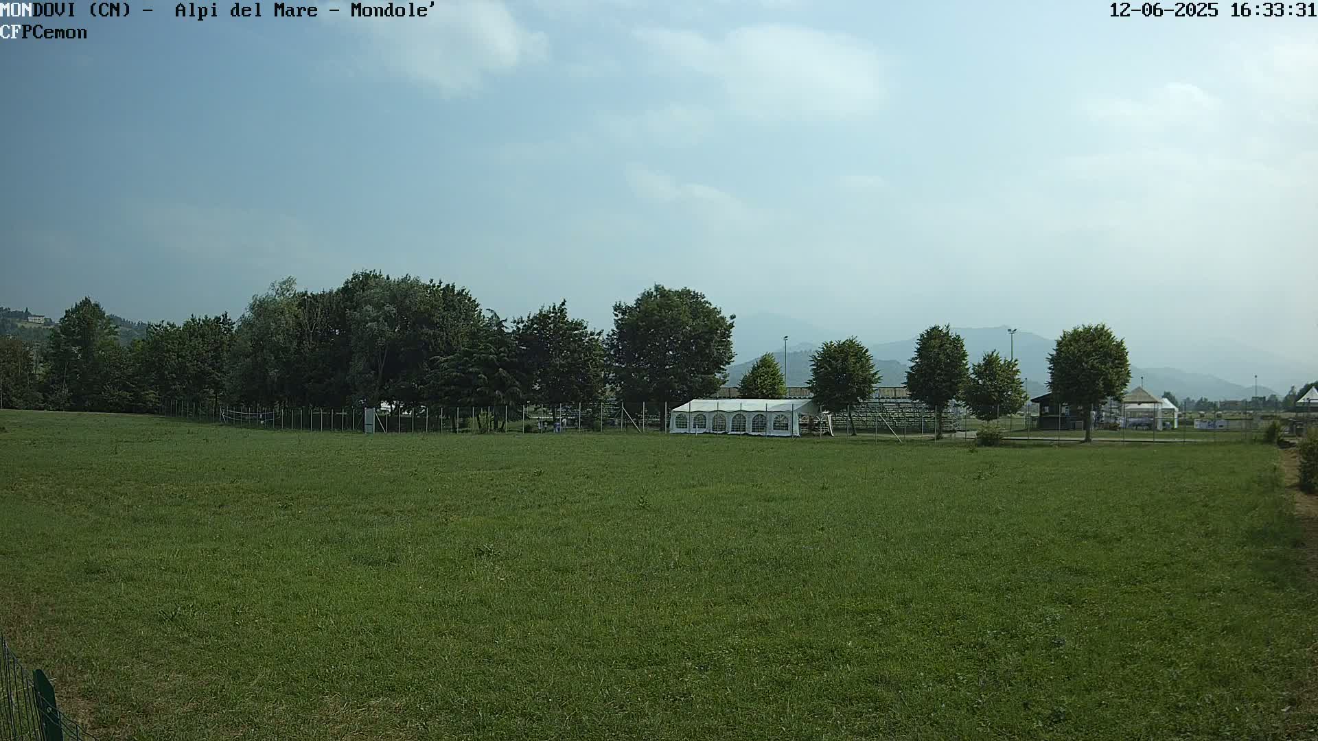 A large, grassy field with a white tent and trees in the background under a partly cloudy sky.