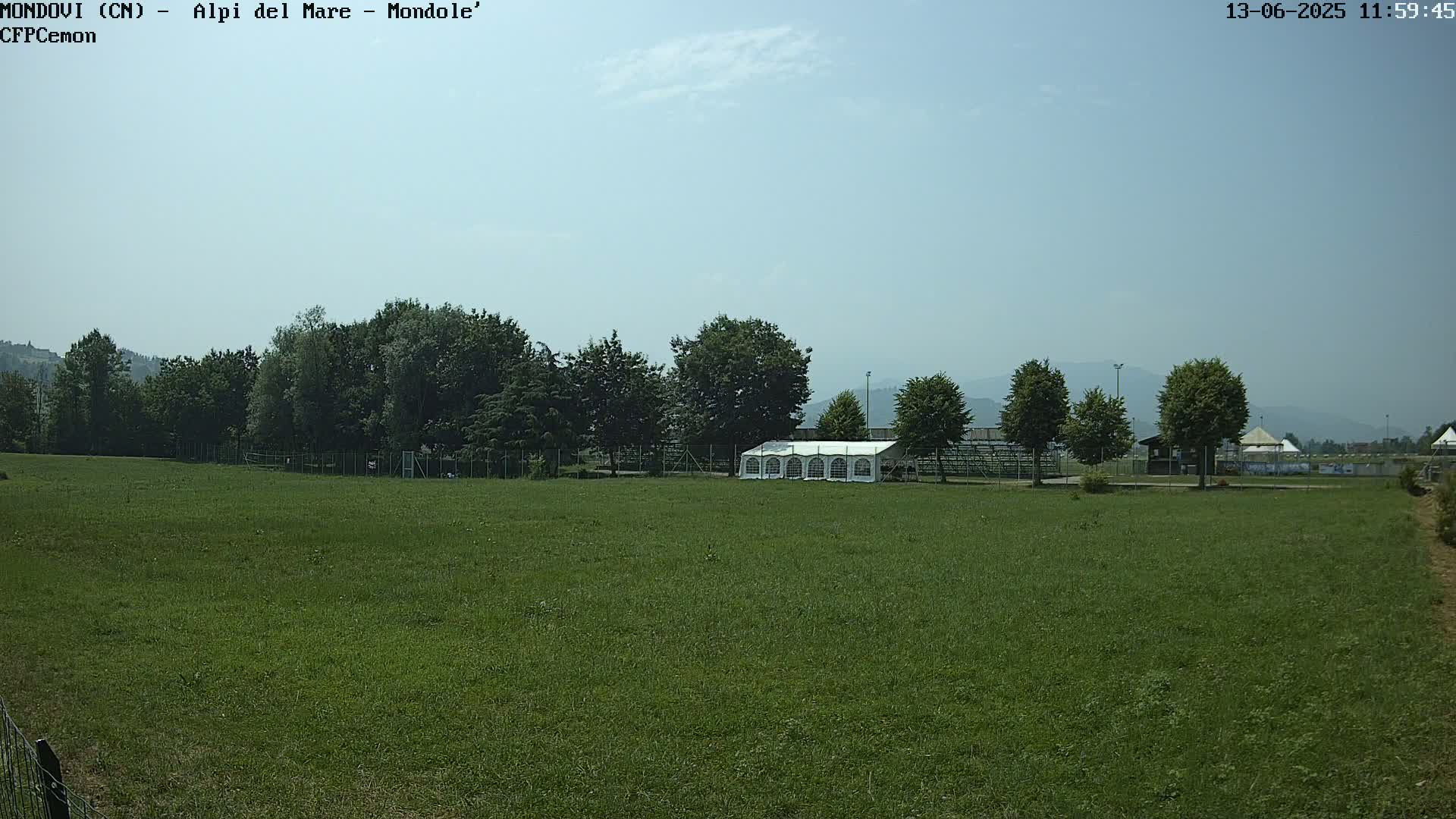 A large grassy field with a white tent sits in the middle of the field, bordered by a line of trees under a mostly clear sky.