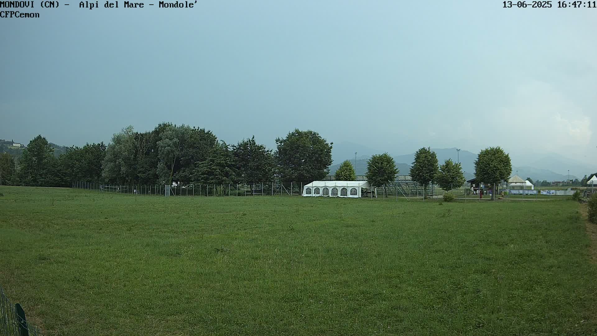 A large grassy field with a white tent, trees, and a fence, under an overcast sky.
