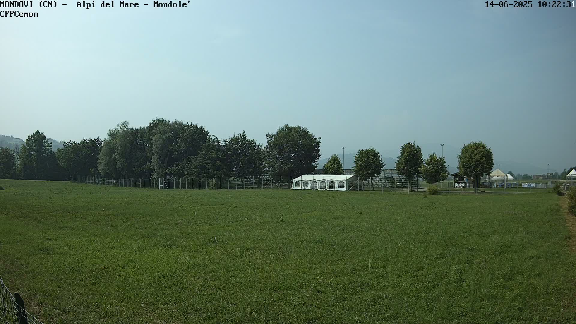 A large grassy field with a white tent and a line of trees in the background under a clear, light-blue sky.