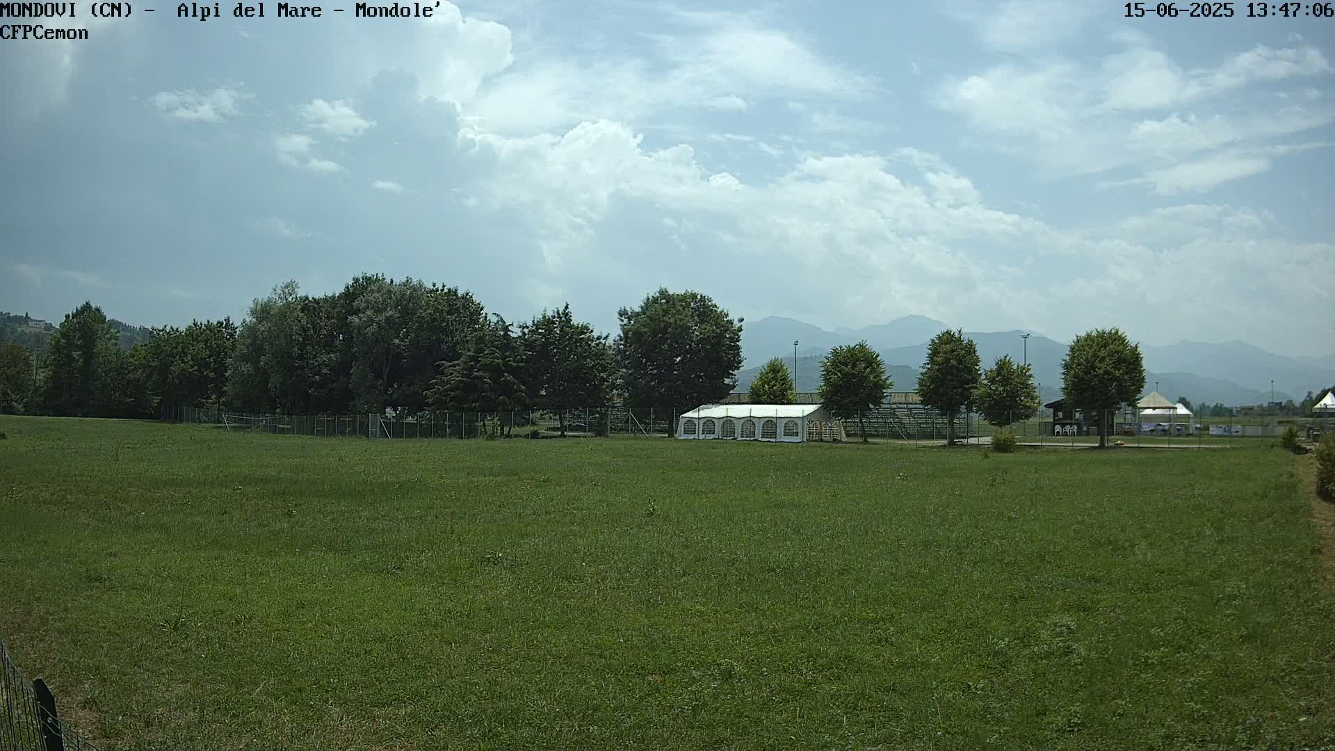 A large grassy field with a white tent, trees, and distant mountains under a partly cloudy sky.