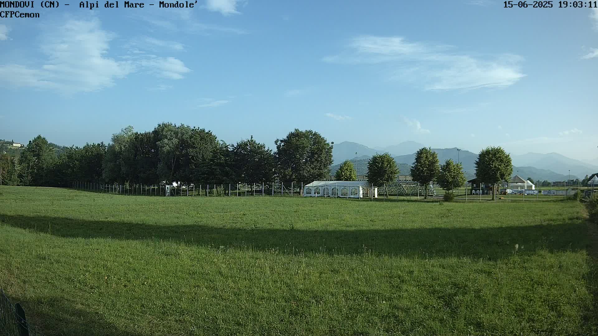 A grassy field stretches towards a line of trees and a white tent, beyond which lie distant mountains under a mostly clear, sunny sky.