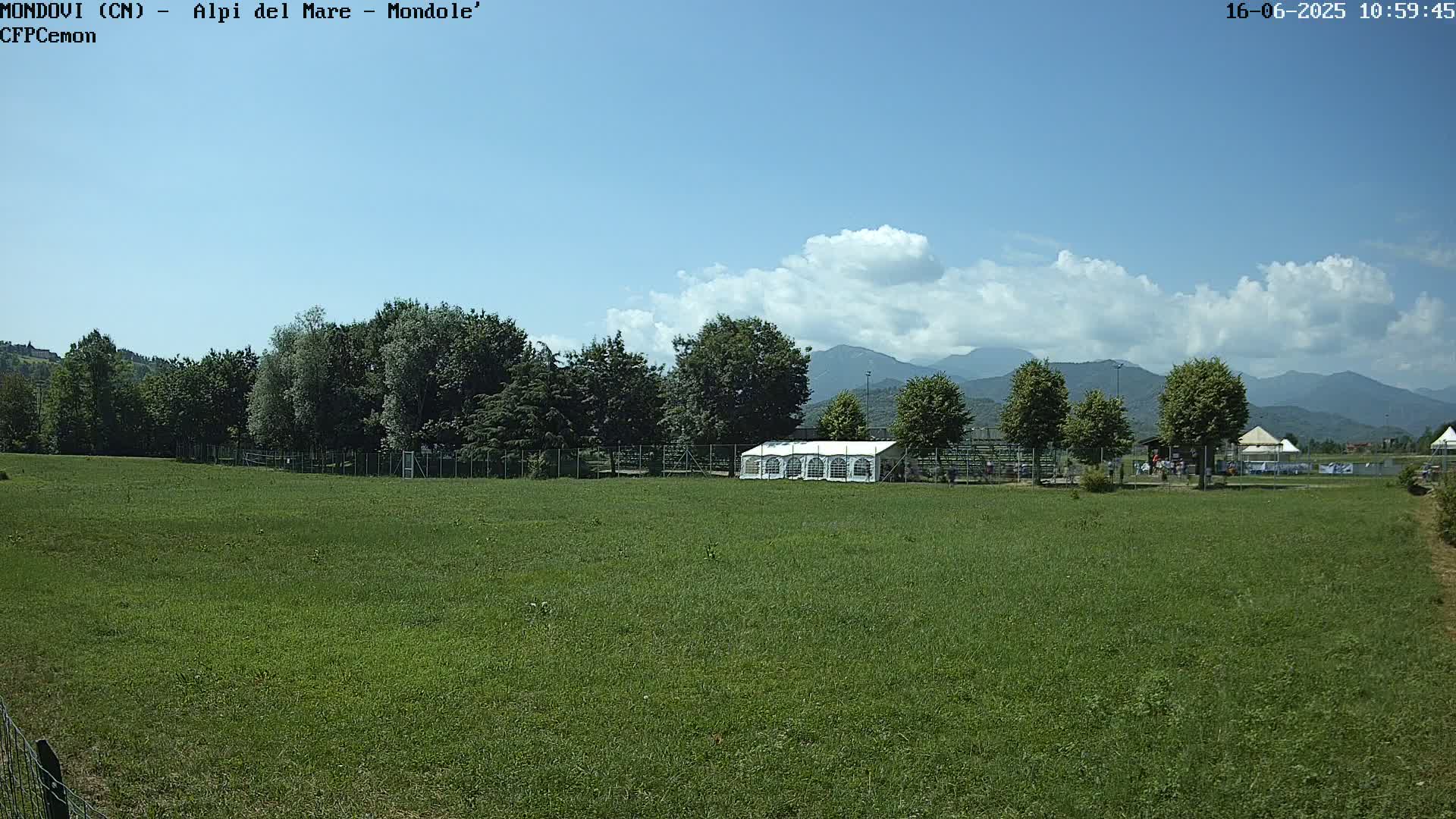 A large grassy field with a white tent and trees in the background, under a partly cloudy blue sky.