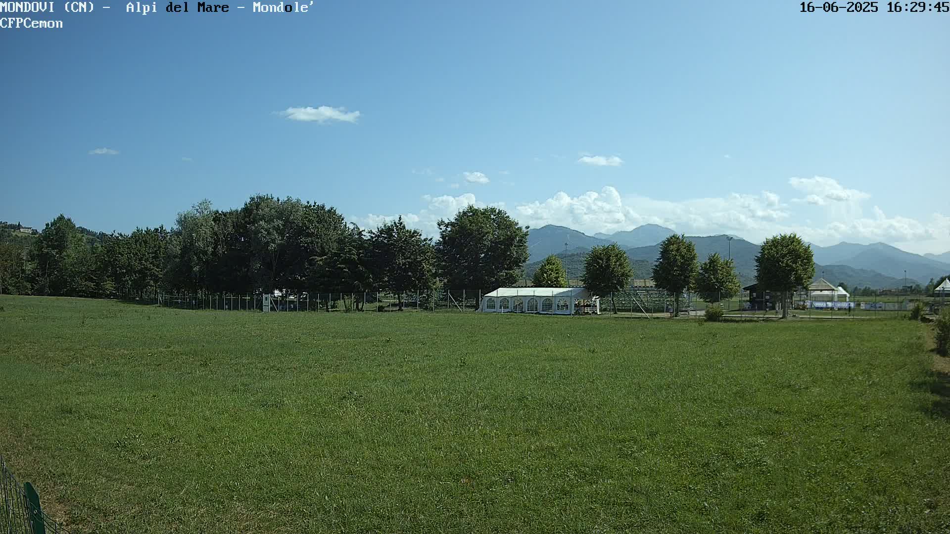 A large grassy field with a white tent and trees in the background, under a mostly sunny sky with some clouds and mountains in the distance.