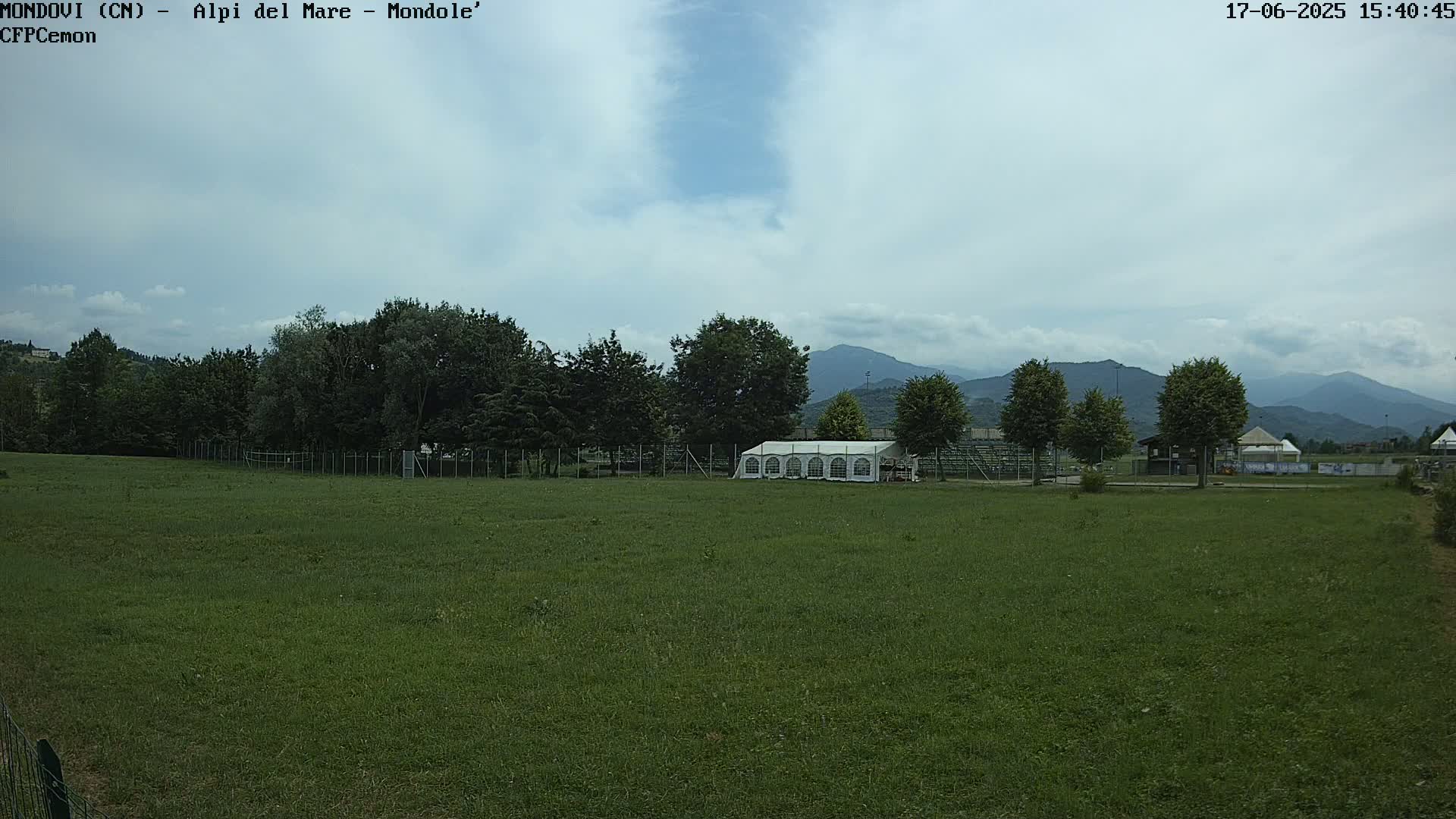 A large grassy field with a white tent and trees in the background, under a mostly cloudy sky and distant mountains.