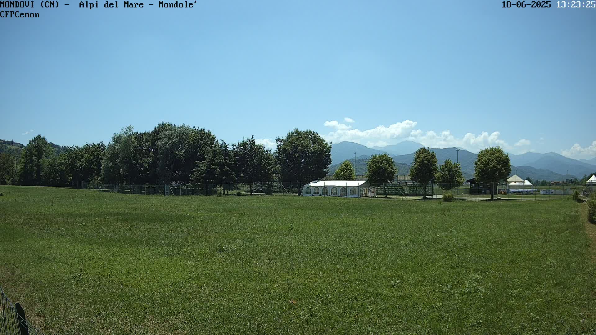 A large grassy field with a white tent and trees in the background, under a mostly clear blue sky with some clouds and distant mountains.