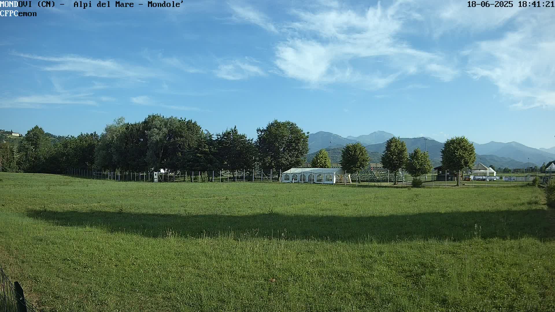 A large grassy field with a white tent and trees in the background under a mostly clear blue sky, mountains visible in the distance.