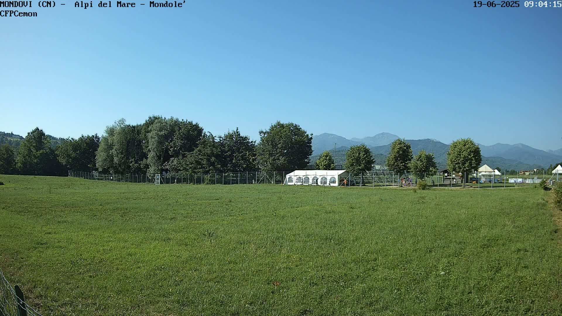 A large grassy field with a white tent and trees sits before a mountain range under a clear blue sky.