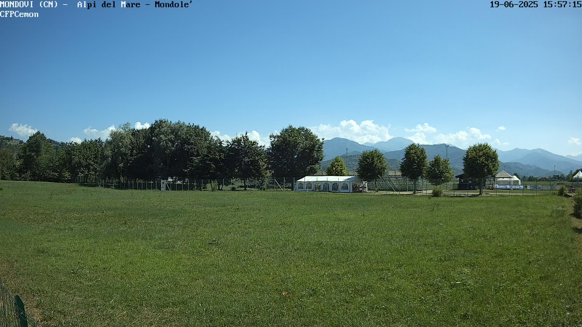 A large grassy field stretches towards a line of trees and a white tent, with mountains in the background under a mostly sunny sky.