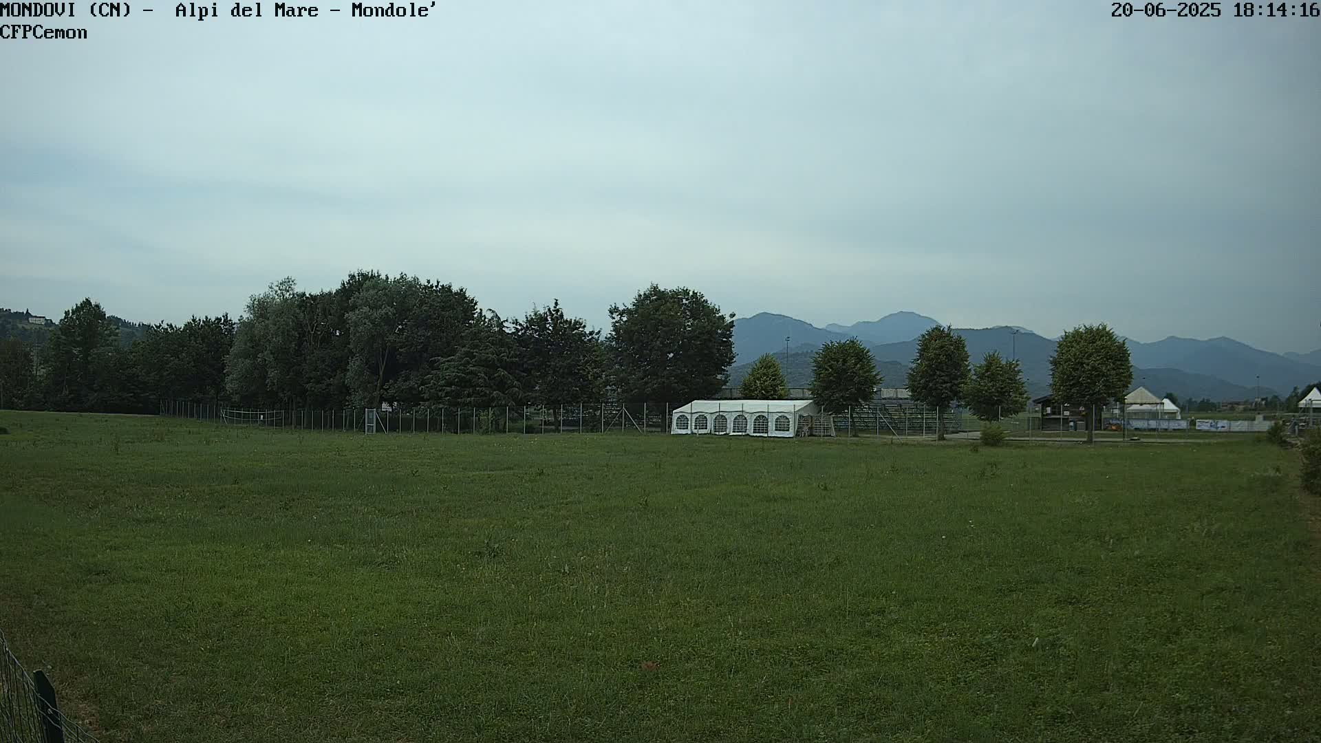 A large grassy field with a white tent and trees behind a fence, under an overcast sky with mountains in the background.