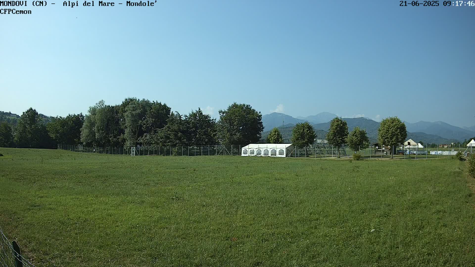 A large grassy field, enclosed by a fence, contains a white tent and is situated in front of a line of trees with mountains in the background under a clear blue sky.