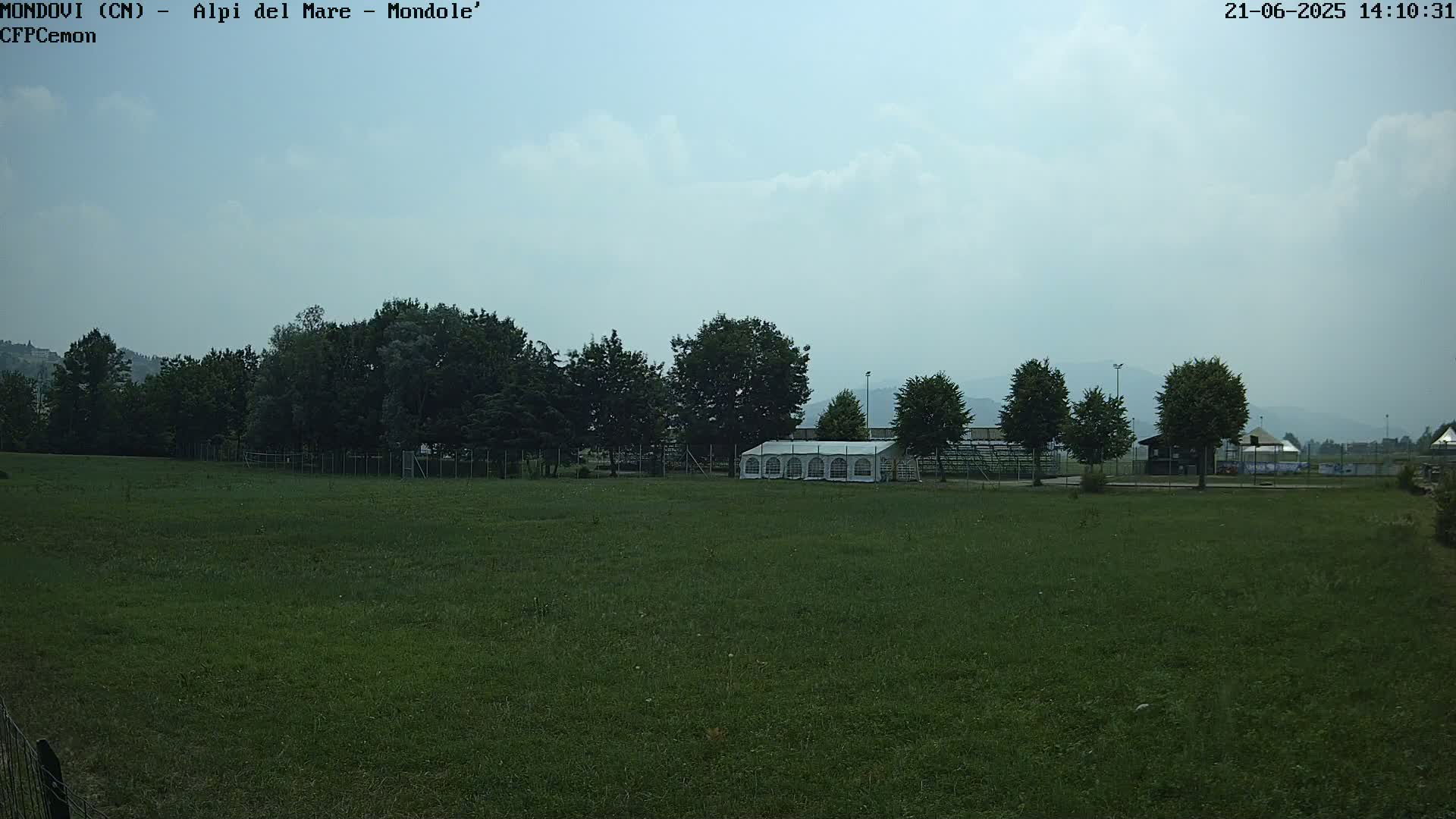 A large grassy field with a white tent and trees behind a fence under a mostly cloudy sky.