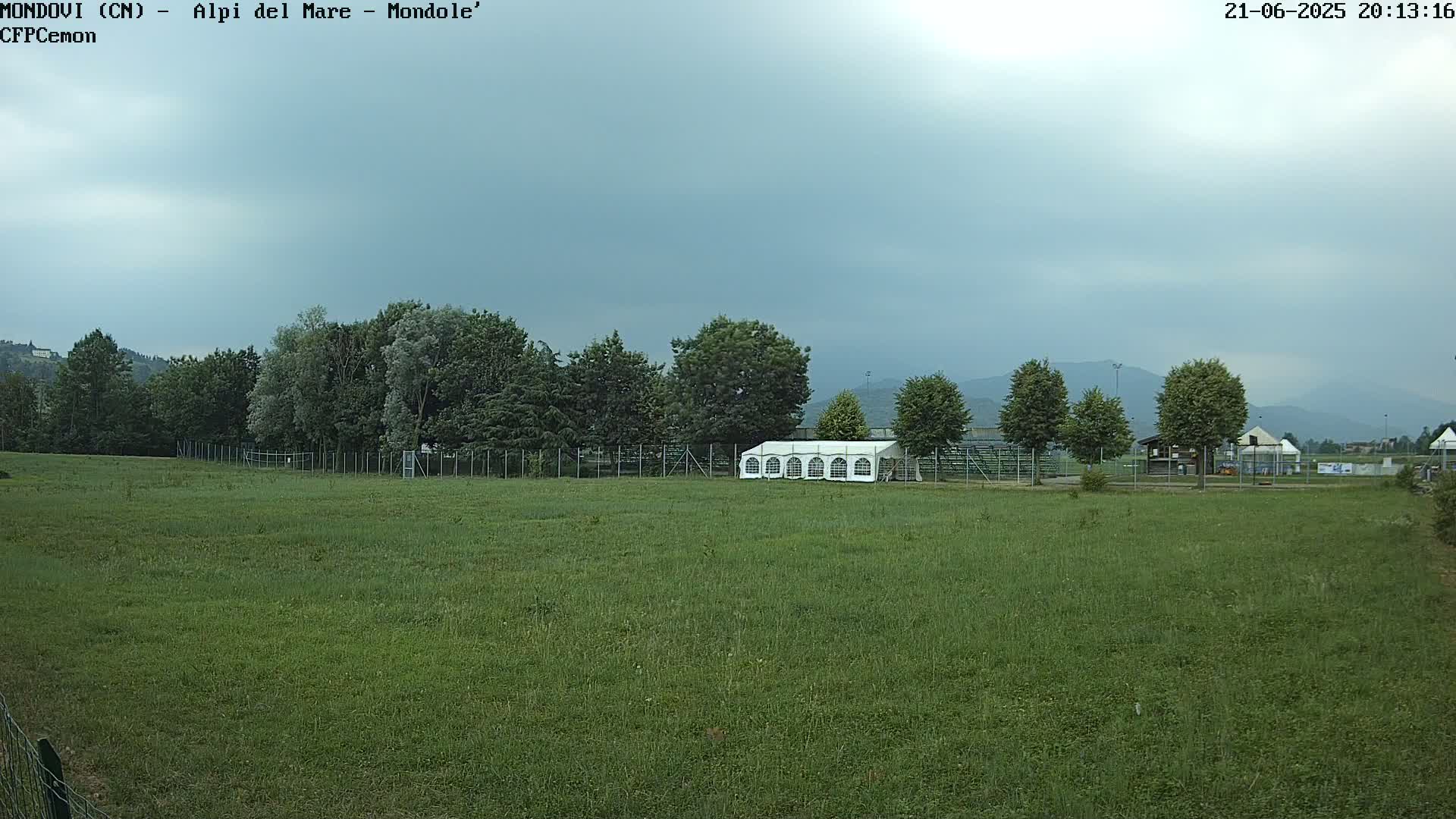 A large, grassy field with a white tent sits behind a fence, in front of a line of trees and distant mountains under an overcast sky.
