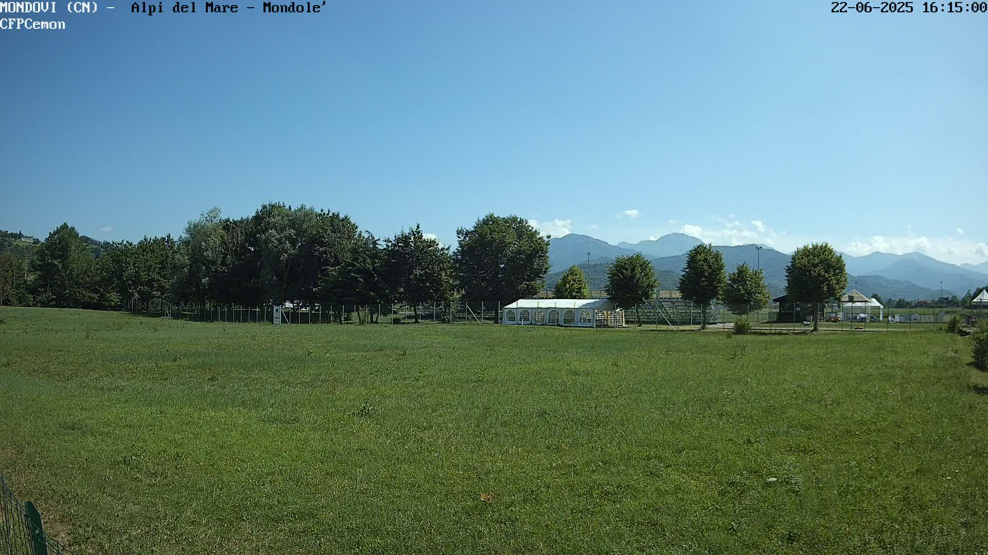 A grassy field with a white tent sits in front of a line of trees, and mountains are visible in the background under a clear blue sky.