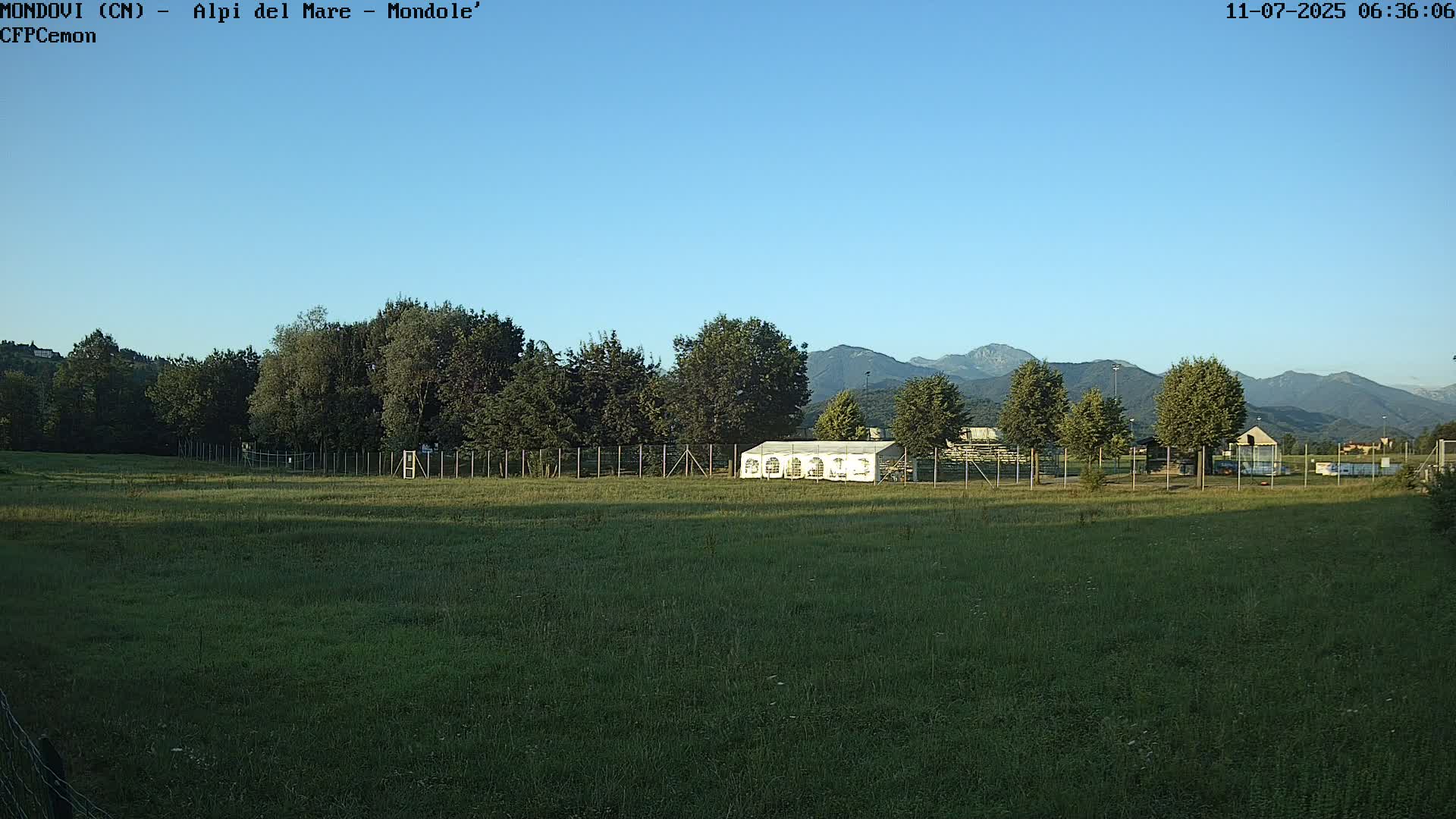 A large grassy field with a white tent and trees in the midground, mountains in the background under a clear blue sky.