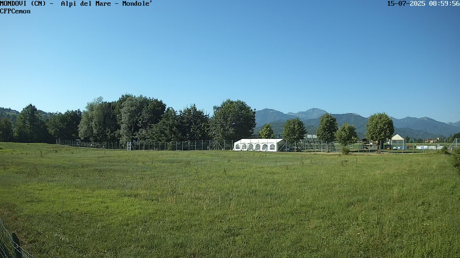 A large grassy field with a white tent sits in front of a line of trees and a distant mountain range under a clear blue sky.