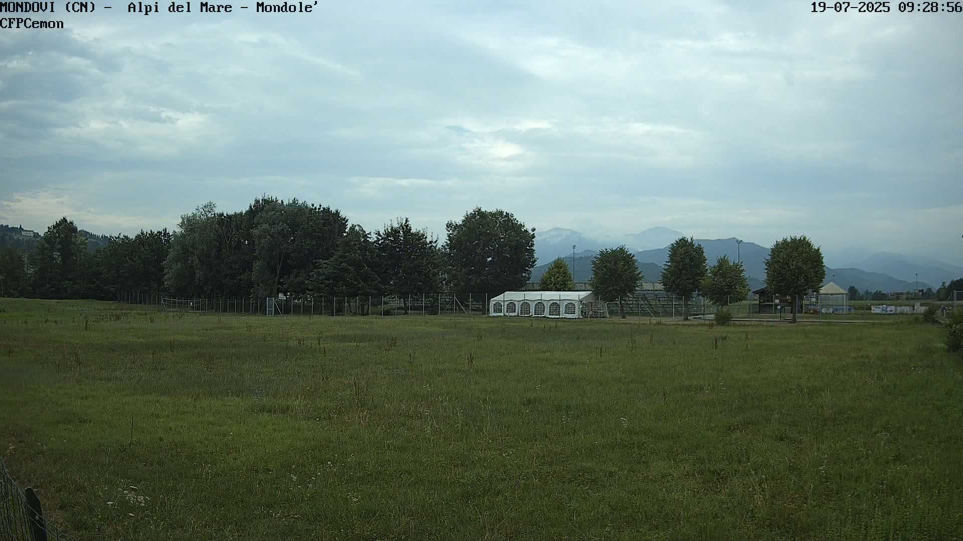 A large grassy field with a white tent and trees in the background under a cloudy sky.