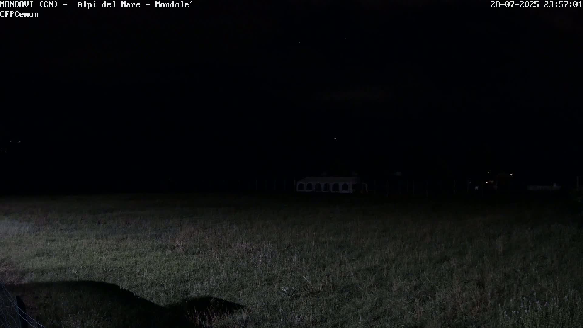 A dark, nighttime scene shows a grassy field with a structure in the distance under a clear sky.