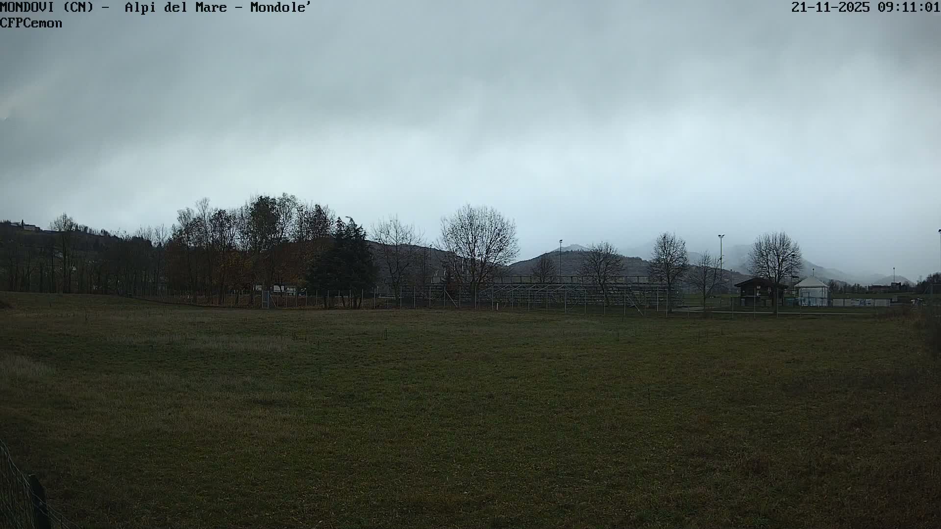 On an overcast and misty day, a wide grassy field leads to a line of mostly bare trees, some bleachers, and a small building, all set against distant, fog-shrouded hills.