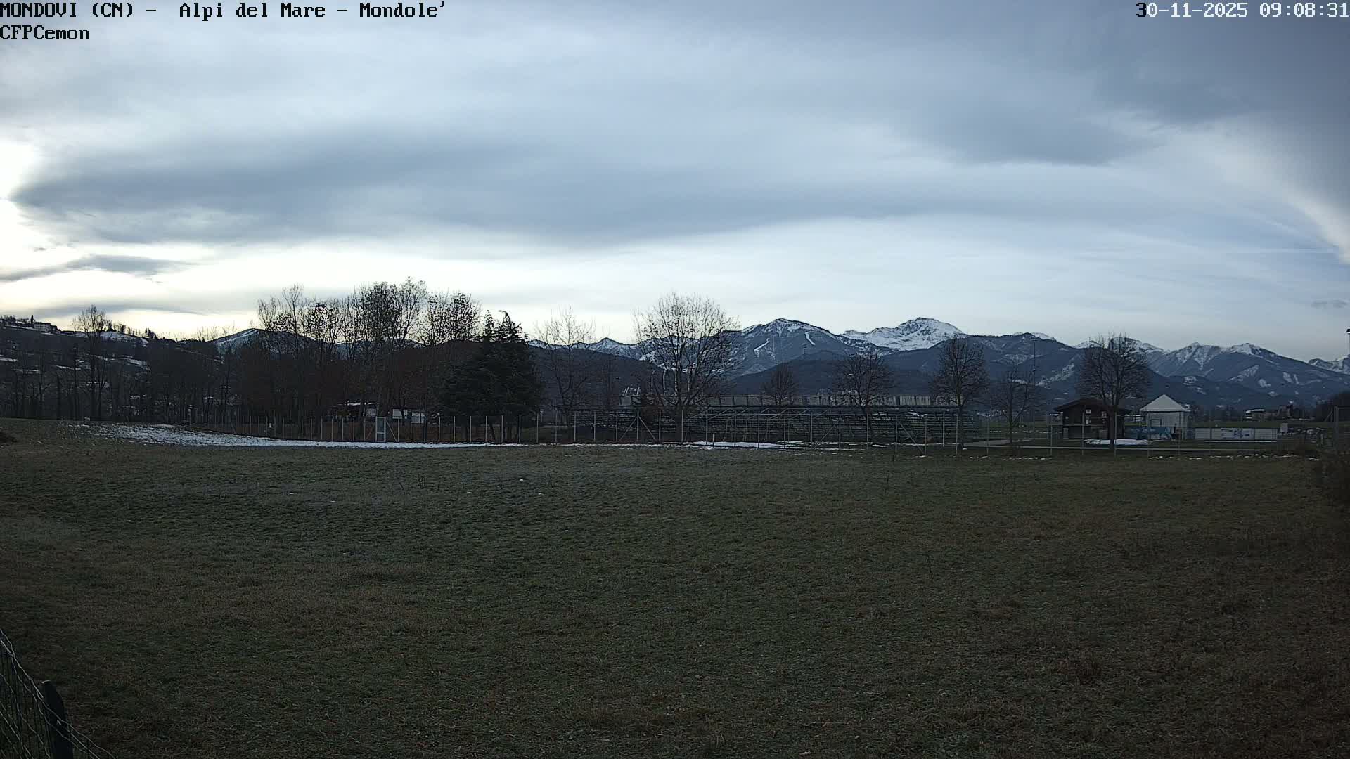 An outdoor panoramic view shows a dormant, slightly frosted field in the foreground, leading to a line of bare trees, fences, and a few small buildings with scattered snow, all set against a backdrop of distant, snow-capped mountains under a heavily overcast sky.