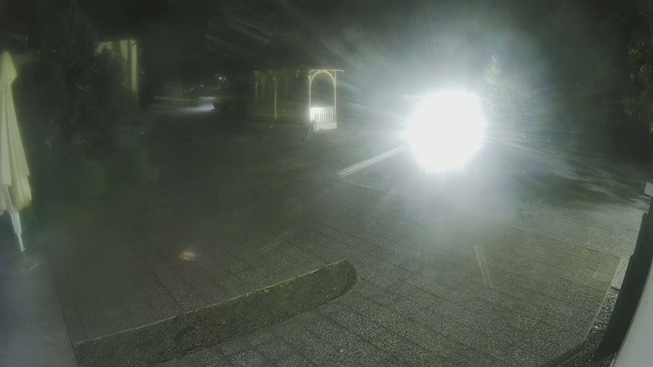 A nighttime view of a gravel driveway with a gazebo and part of a patio umbrella visible, illuminated by bright headlights.