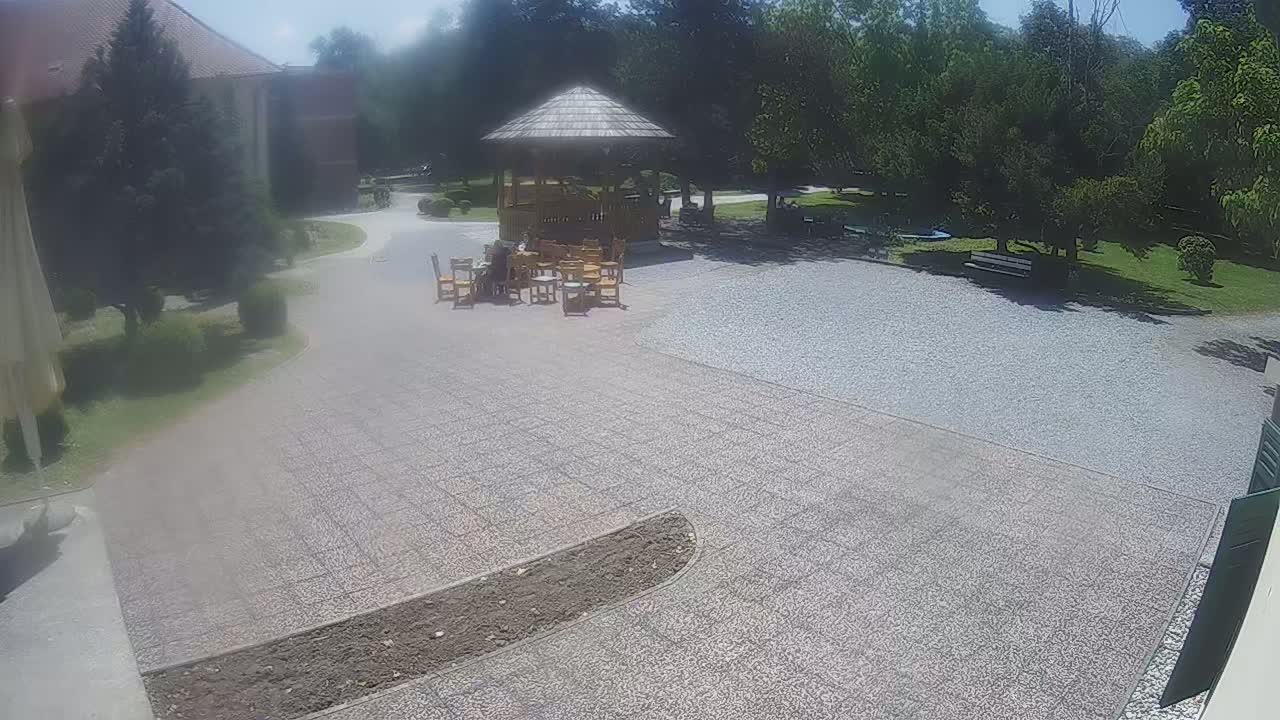 A gravel courtyard with a wooden gazebo and several people sitting at tables, under a sunny sky.
