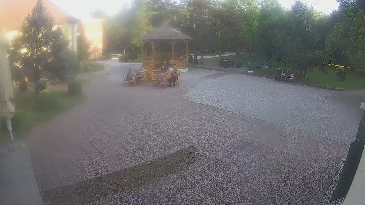 A group of people are sitting at tables outside a wooden gazebo in a courtyard on a hazy, sunny day, with other people sitting on a bench nearby.