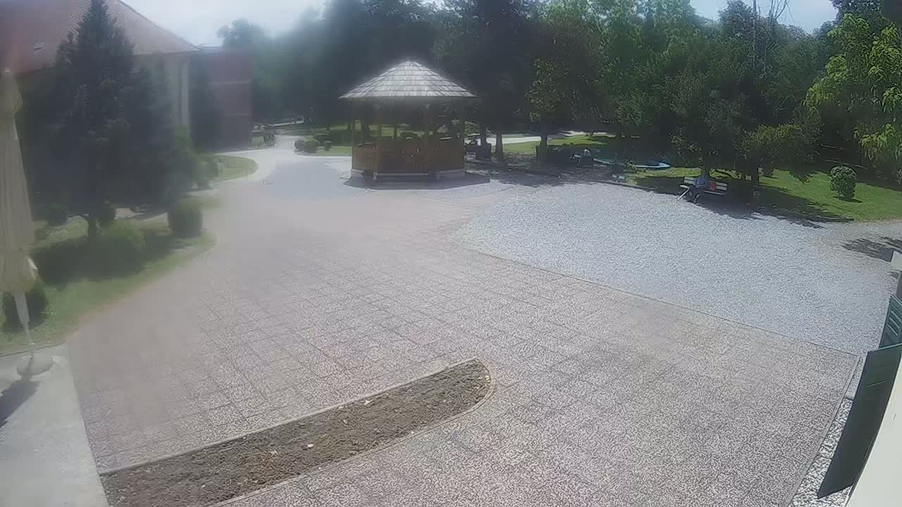A gravel courtyard with a wooden gazebo, surrounded by trees and a building on a sunny day.