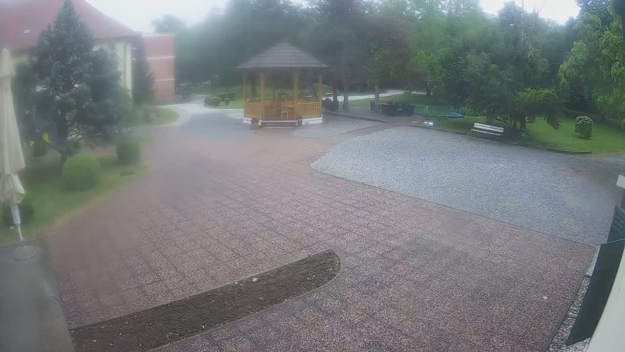 A rain-soaked gravel courtyard features a wooden gazebo, a bench, and landscaping, with a building visible in the background.