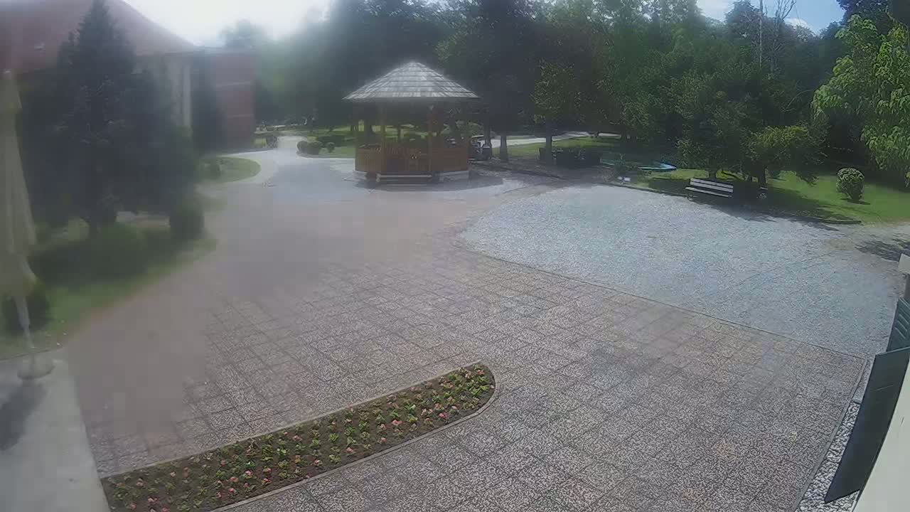 A paved area with a wooden gazebo, flowerbeds, a park bench, and trees, under a partly sunny sky.