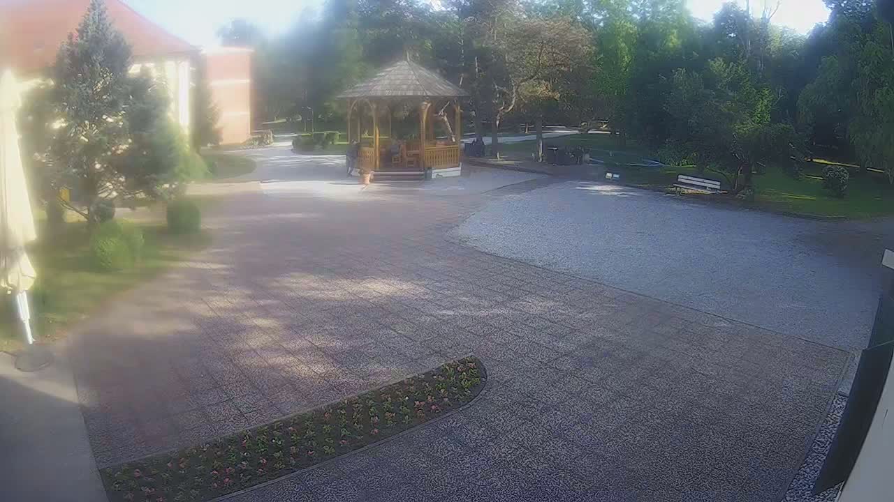 A paved courtyard with a wooden gazebo, surrounded by trees and landscaping, on a sunny day.