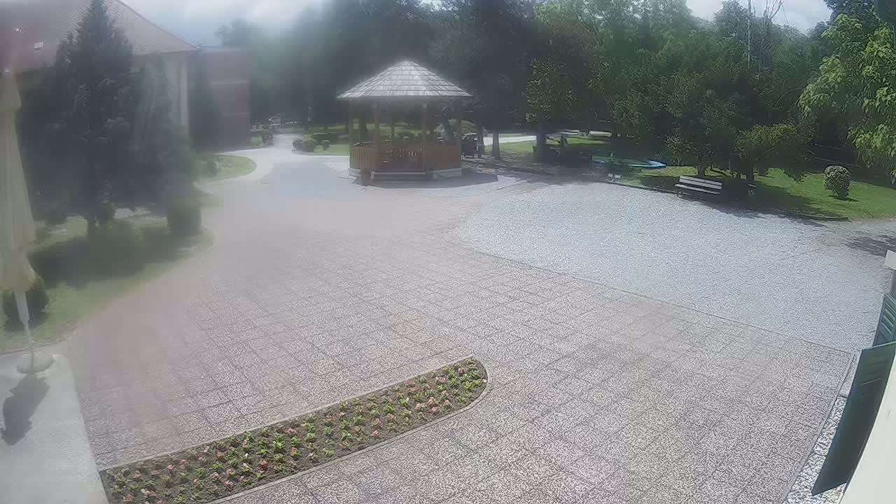 A paved courtyard with a wooden gazebo, flowerbeds, park benches, and trees under a partly cloudy sky.