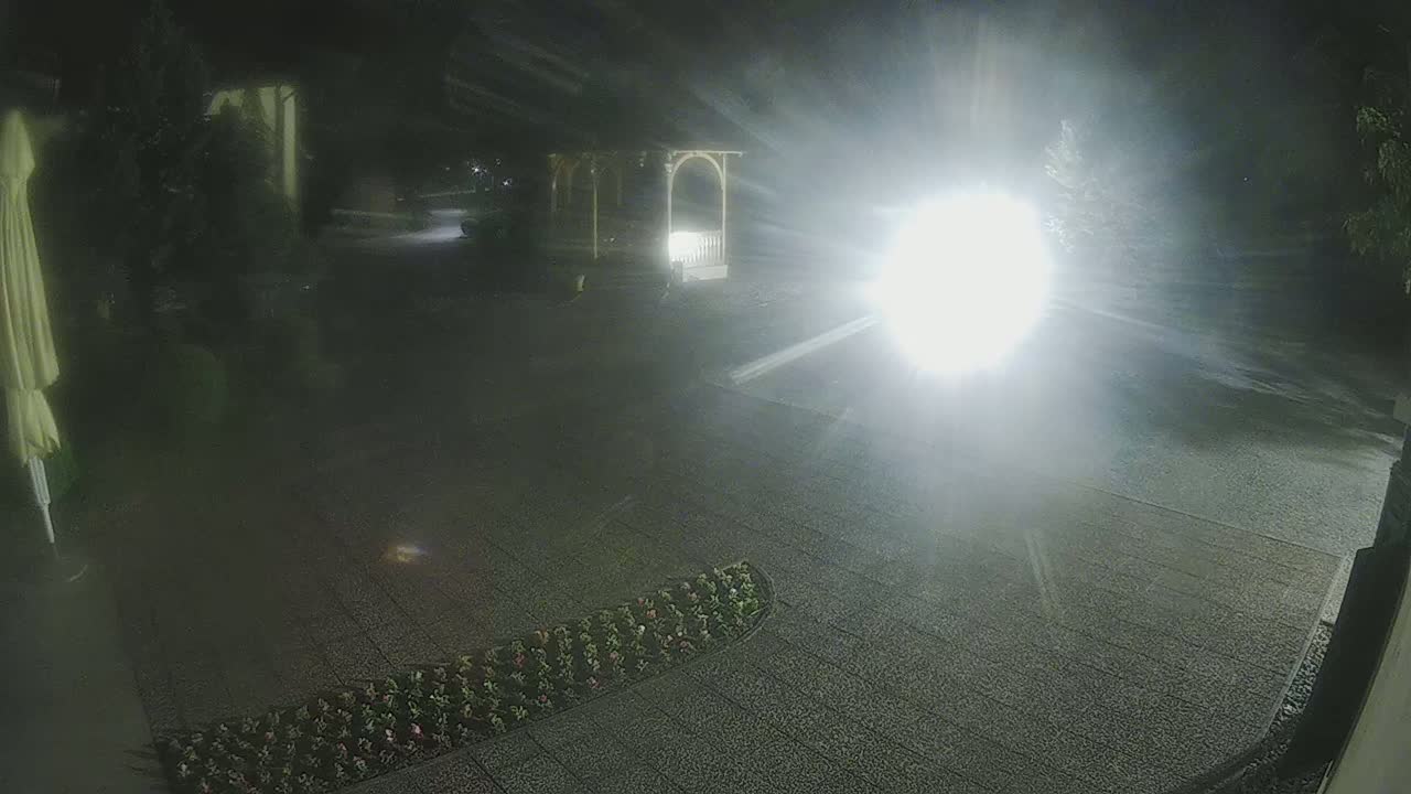 A nighttime outdoor scene shows a paved area with a gazebo and flowerbed, illuminated by a bright light source.