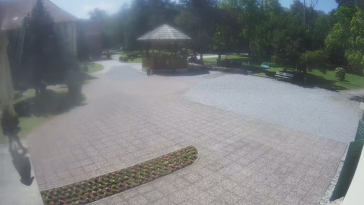 A gravel courtyard with a wooden gazebo, a flower bed, and a park-like setting under a sunny sky.