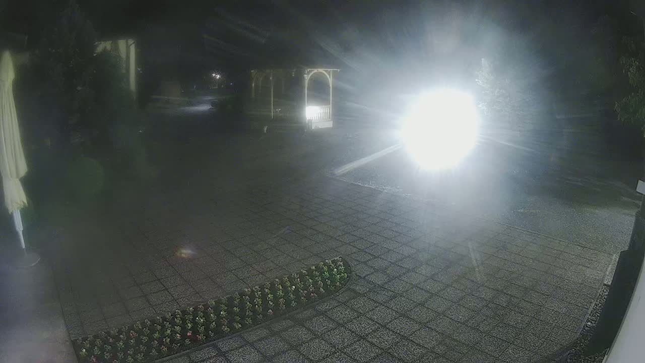 A nighttime view of a paved area with a gazebo and a bright light source creating glare, with low-lying vegetation bordering a section of the pavement.