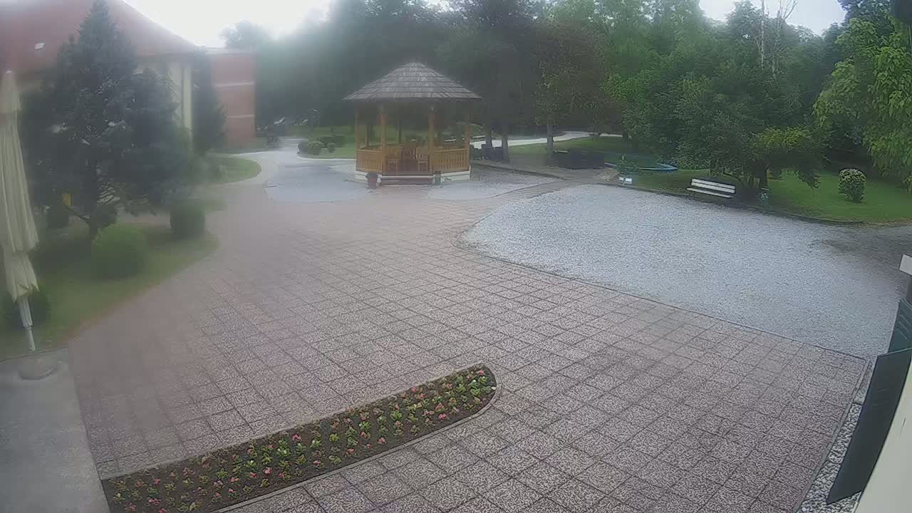 A paved courtyard with a wooden gazebo, surrounded by trees and a flower bed, on an overcast day.
