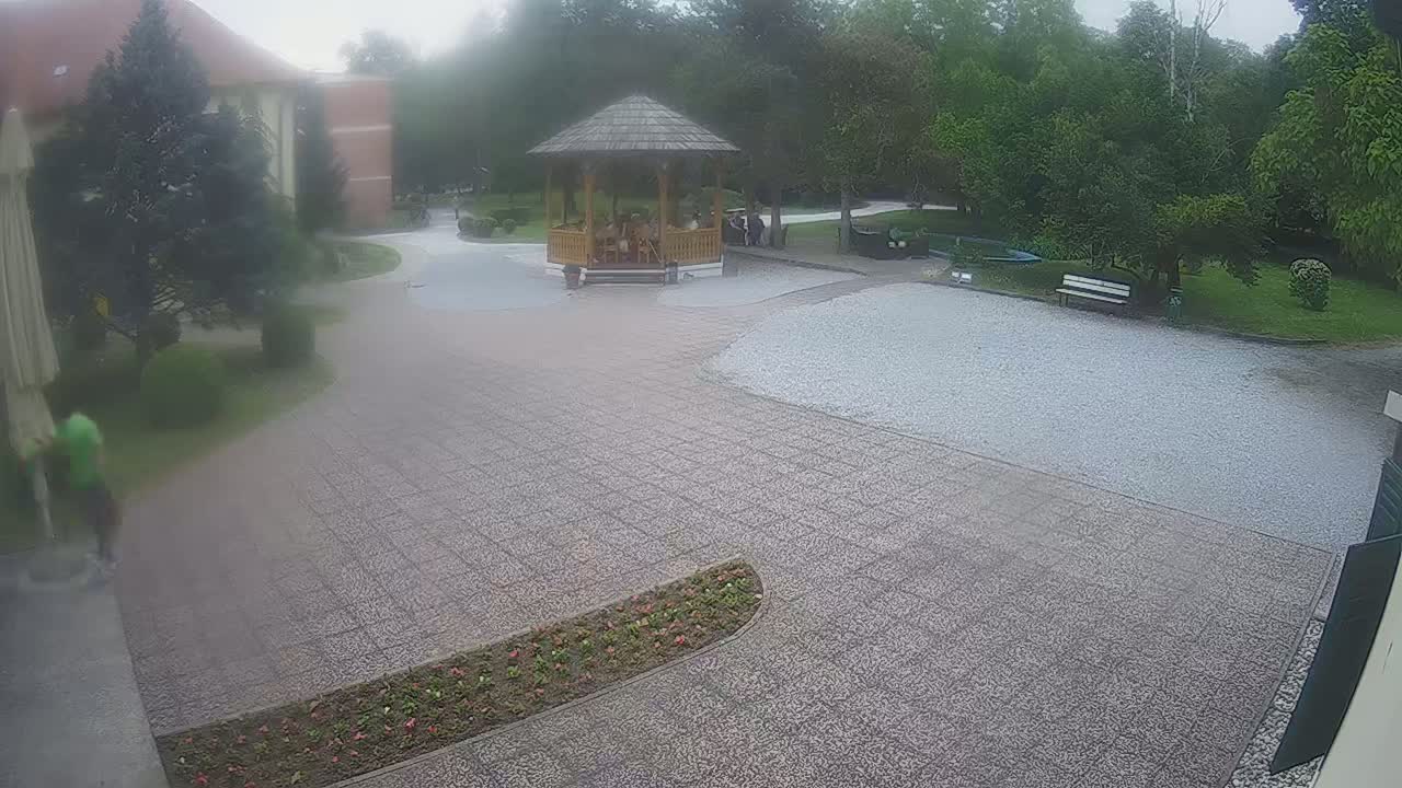 A paved courtyard with a gazebo containing several people, surrounded by trees and landscaping, on an overcast day.