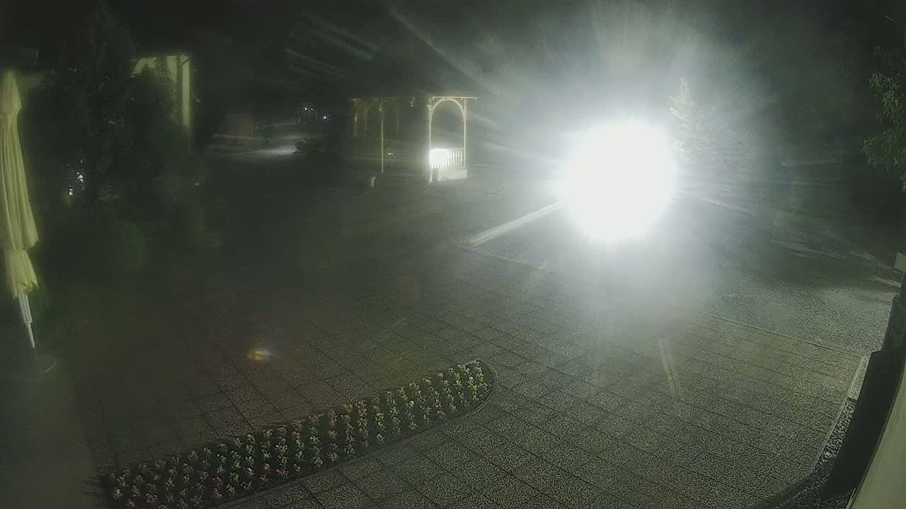 A nighttime outdoor scene shows a paved area with a flowerbed, a gazebo, and a bright light source creating glare.