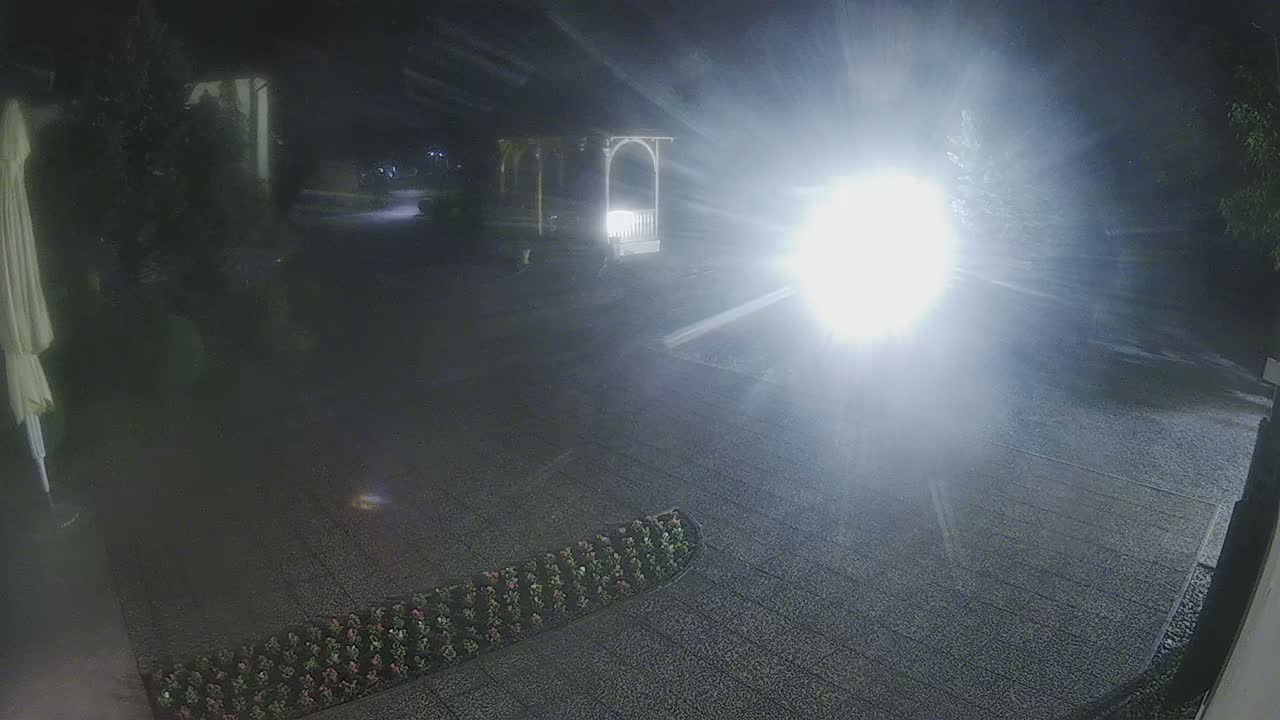 A nighttime view of a gravel area with a gazebo, some low-lying plants, and a bright light source creating glare.