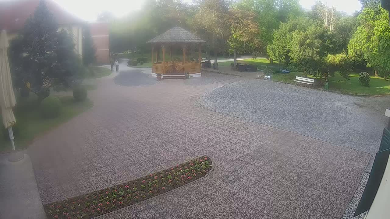 A hazy outdoor scene shows a paved courtyard with a wooden gazebo, some landscaping, and a few people walking in the distance.