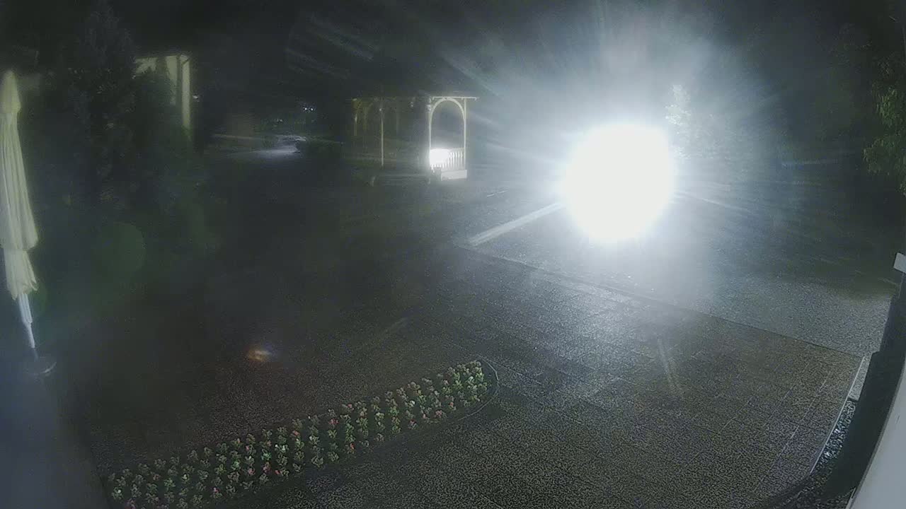 A nighttime, rain-slicked driveway is shown with a gazebo and a row of flowers in the foreground, illuminated by bright headlights approaching from the middle distance.